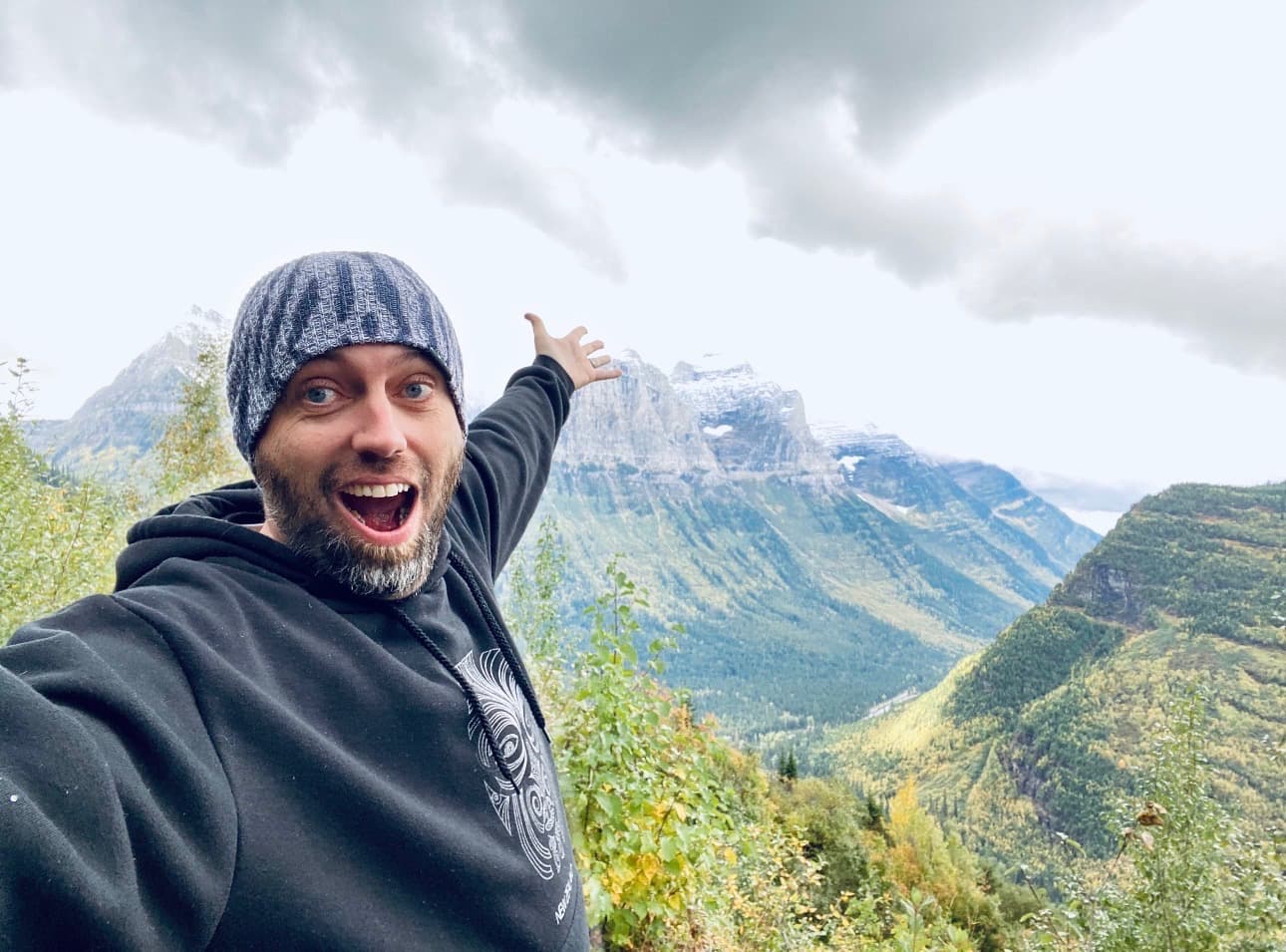 Marc Kennedy in a beanie and hoodie, pointing back at dramatic mountain peaks behind him