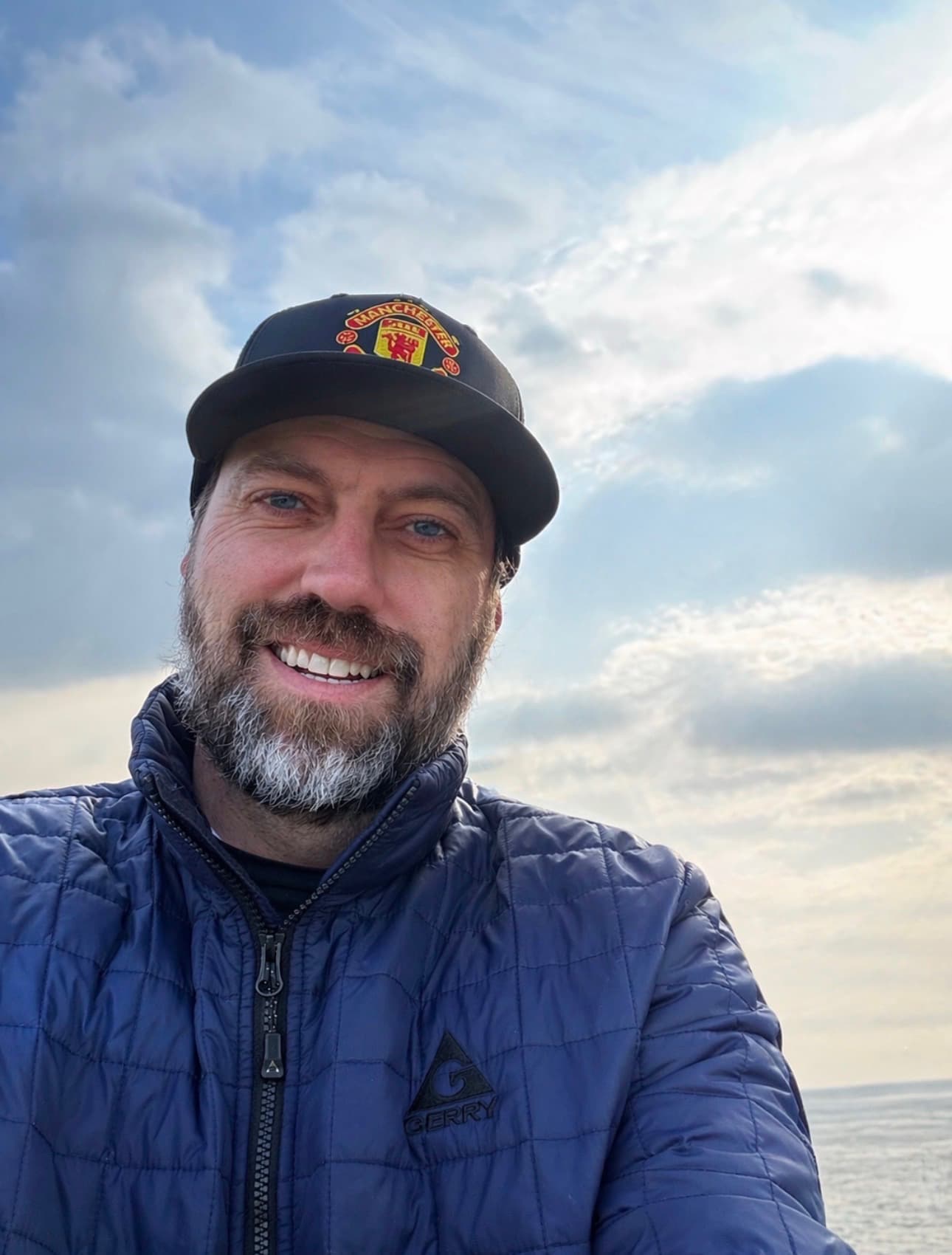 Marc Kennedy wearing a Manchester United cap and a navy Gerry puffer jacket, ocean horizon behind him